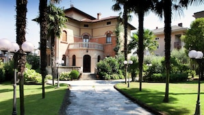 A stone pathway leading to a building with a balcony and arched windows. - Elegant Art Nouveau Villa With Private Pool Near the Lake (Desenzano del Garda)