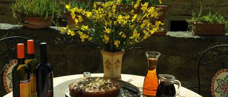 Una mesa redonda con un pastel, botellas de vino, una jarrón y plantas en macetas con flores amarillas y rosas.