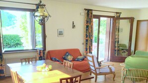 A dining area with a wooden table, chairs, and a fruit bowl. There is a sofa with cushions and a chair with a cushion. A window provides natural light, and a door leads to an outdoor area. - Villa in Northern Italy Near Borgo di Toppo (Travesio)