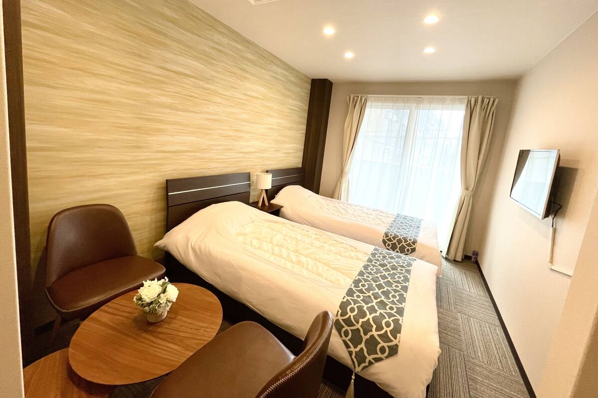 A hotel room with two beds, a desk, a chair, a TV, and a window with curtains.