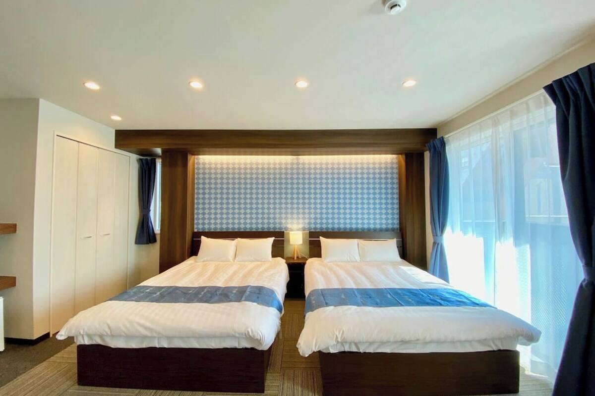 A hotel room with two beds, a large window with curtains, a ceiling with recessed lighting, and a wall-mounted security camera.