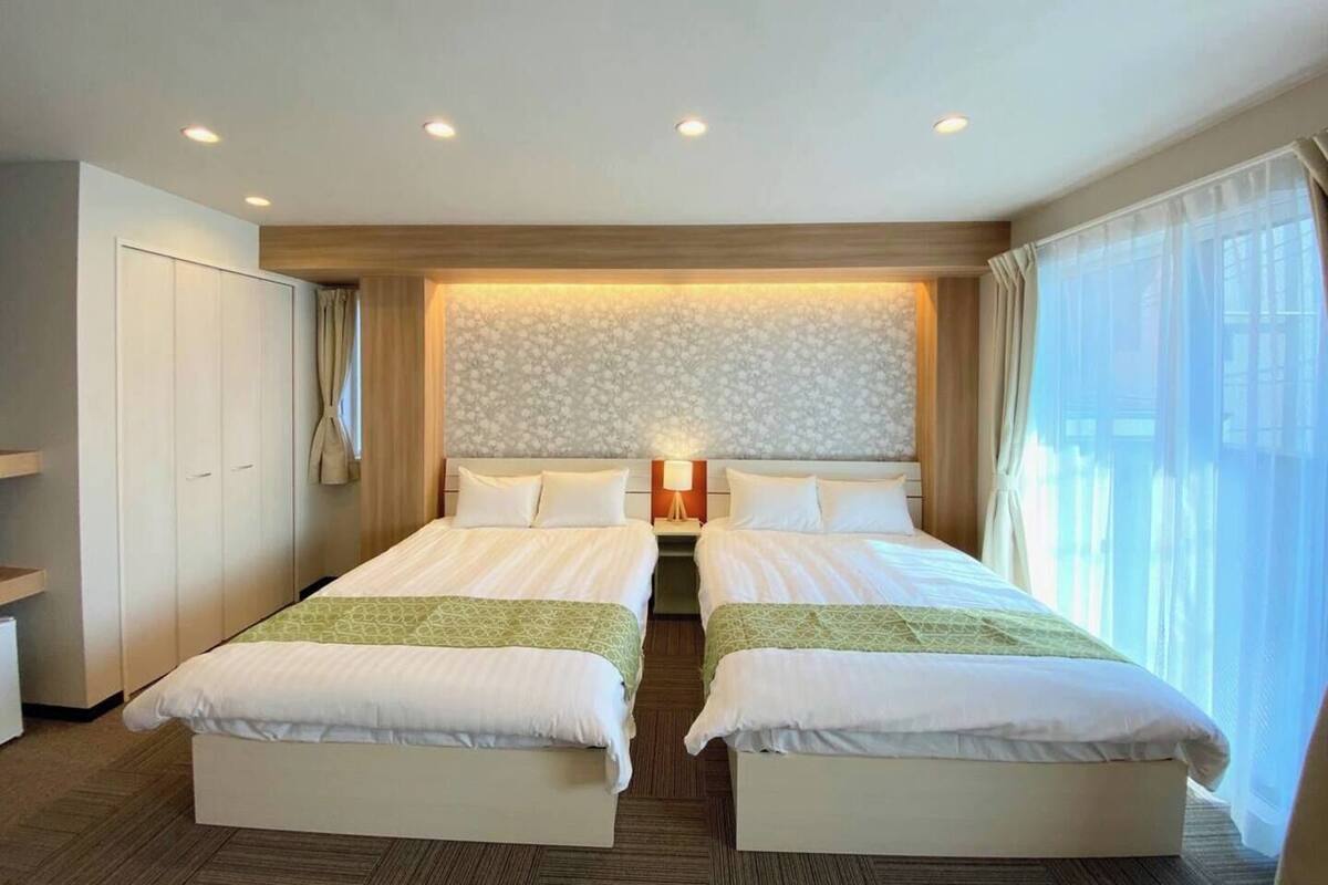 A hotel room with two beds, a wardrobe, a nightstand with a lamp, and a large window with curtains.
