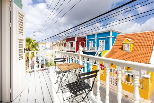 A balcony with a wooden table and black chairs, overlooking colorful buildings with red and blue facades.