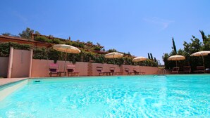Ein Schwimmbecken mit klarem Wasser, umgeben von Liegestühlen und Sonnenschirmen, und eine Backsteinmauer mit Grünpflanzen.