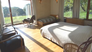 Une chambre avec un grand lit, un fauteuil à bascule, une petite table et une vue sur l’extérieur.