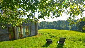 Une maison moderne dotée d’une grande baie vitrée, de deux fauteuils confortables placés sur une pelouse, et d’une table en bois.