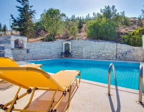 Una piscina con agua azul cristalina, rodeada de muros de piedra y árboles. Se observan sillas de sol amarillas y una pequeña construcción de piedra con una puerta.