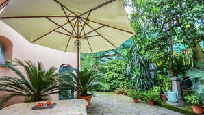 Een met stenen bestraat binnenterrein met een tafel en stoelen onder een grote parasol, omringd door weelderige begroeiing en planten in potten.