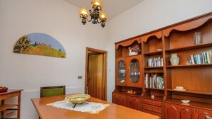 Un comedor con una mesa de madera, sillas, una araña, una estantería y un cuadro en la pared.