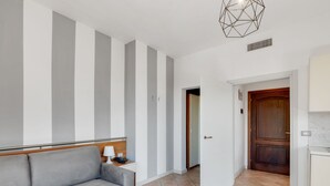Una habitación de hotel moderna con un sofá gris, un cabecero de madera, una lámpara de diseño geométrico y una zona de cocina con una puerta de madera.