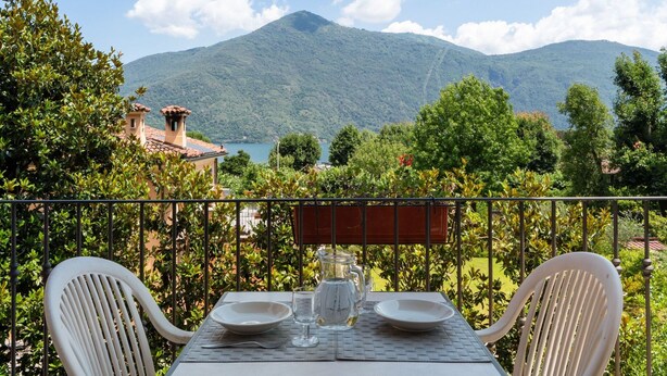 Una mesa con dos sillas blancas, una jarra de vidrio y dos platos preparados para una comida. La mesa está al aire libre, con vista a una montaña y un lago.