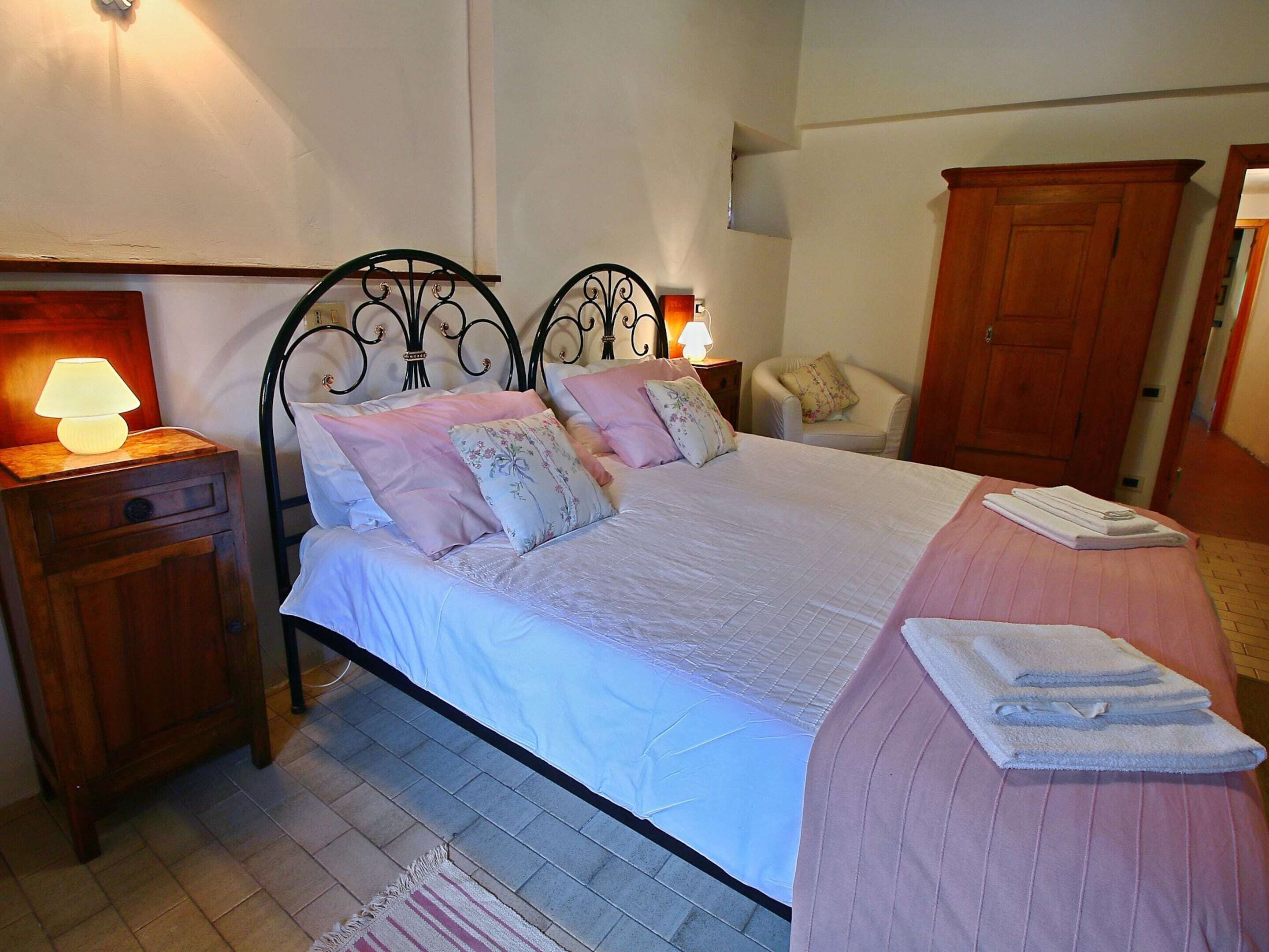 A neatly made bed with a pink bedspread and multiple pillows, a wooden nightstand with a lamp, a chair with a floral cushion, and a wooden wardrobe.