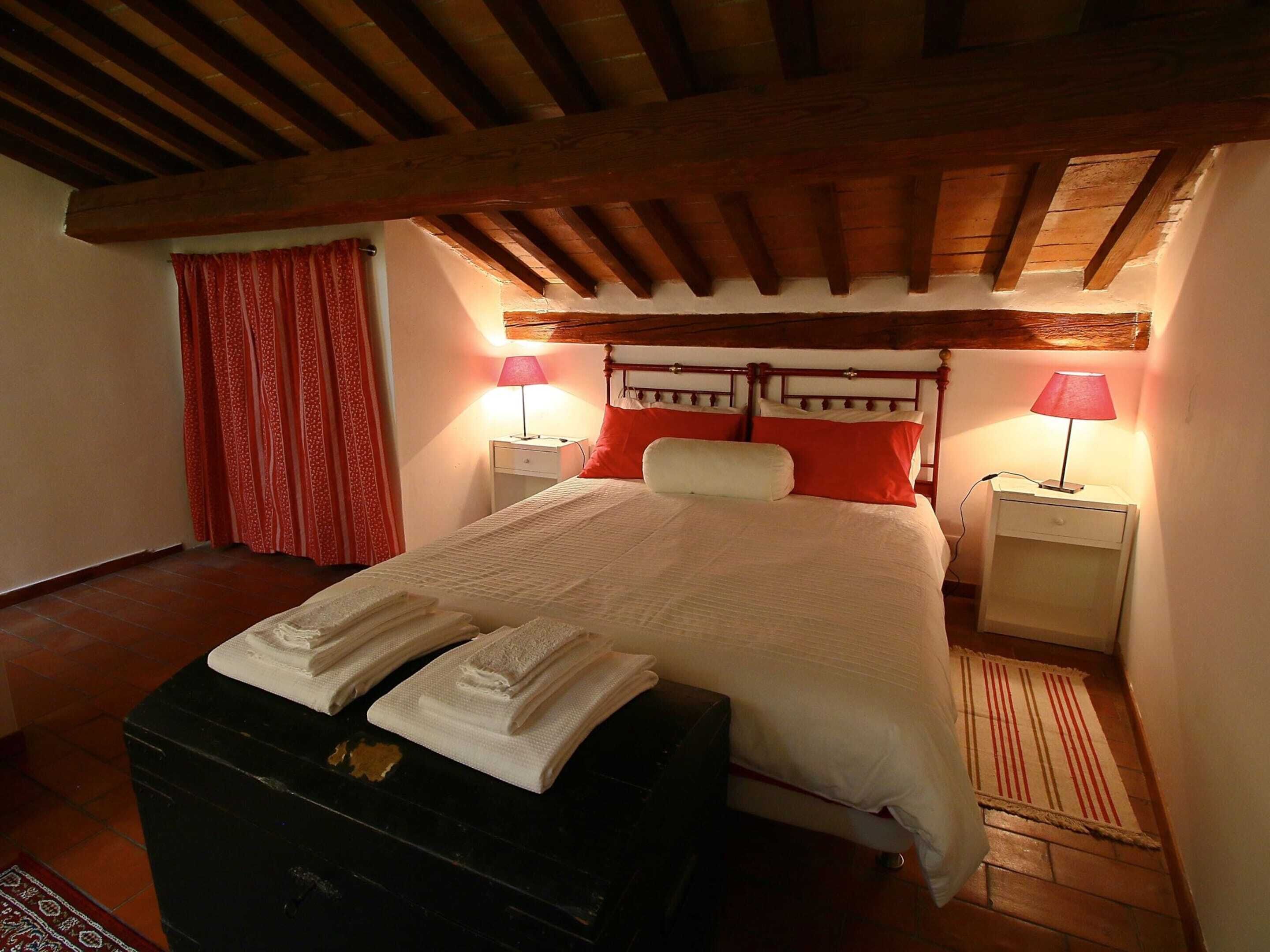 Ein Schlafzimmer mit Holzboden, einem Bett mit weißer Bettwäsche, roten Kissen, zwei Nachttischlampen und einer schwarzen Kommode mit Handtüchern.
