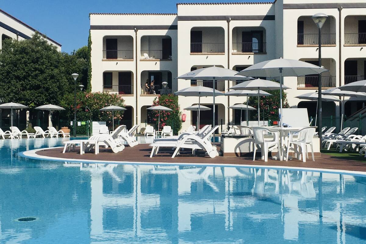 A swimming pool with lounge chairs and umbrellas, surrounded by a building with balconies.
