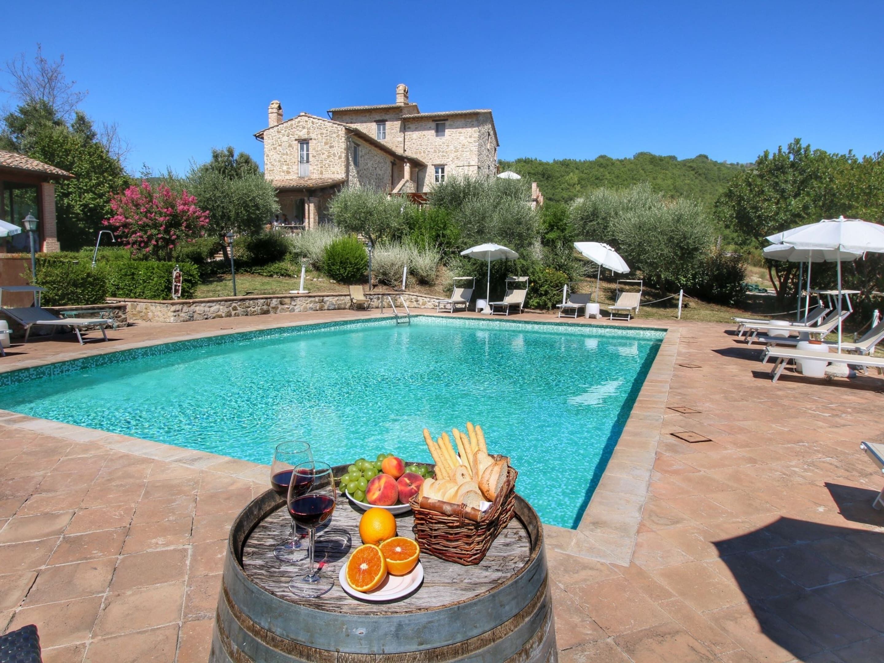 Una piscina con lettini e ombrelloni, un edificio in pietra sullo sfondo e un tavolo con frutta e bicchieri di vino.
