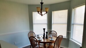 A dining room with a round wooden table, four wooden chairs, a chandelier, and two windows with blinds.