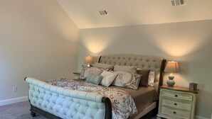 Un dormitorio con cama y cabecera capitonada, ropa de cama estampada, dos mesitas de noche con lámparas y suelo cubierto de alfombra.