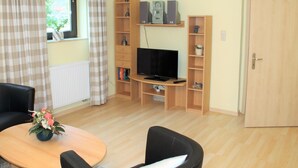 A living room with a TV, wooden shelves, a door, and a window with curtains. - Ferienwohnung, Nienhagen (Nienhagen)