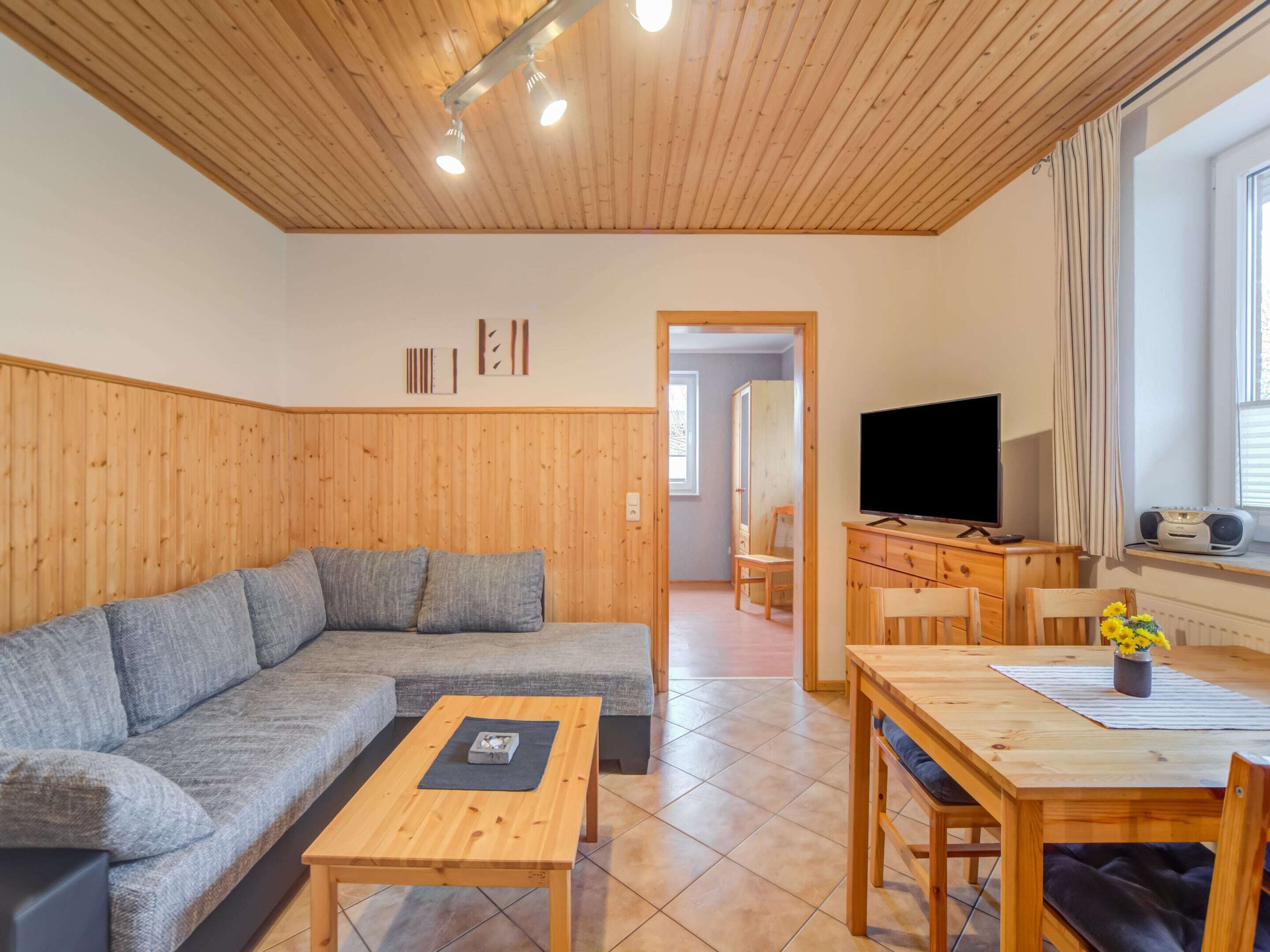 A cozy living room with a wooden ceiling, a sofa, a coffee table, a TV, and a dining area with a table and chairs.