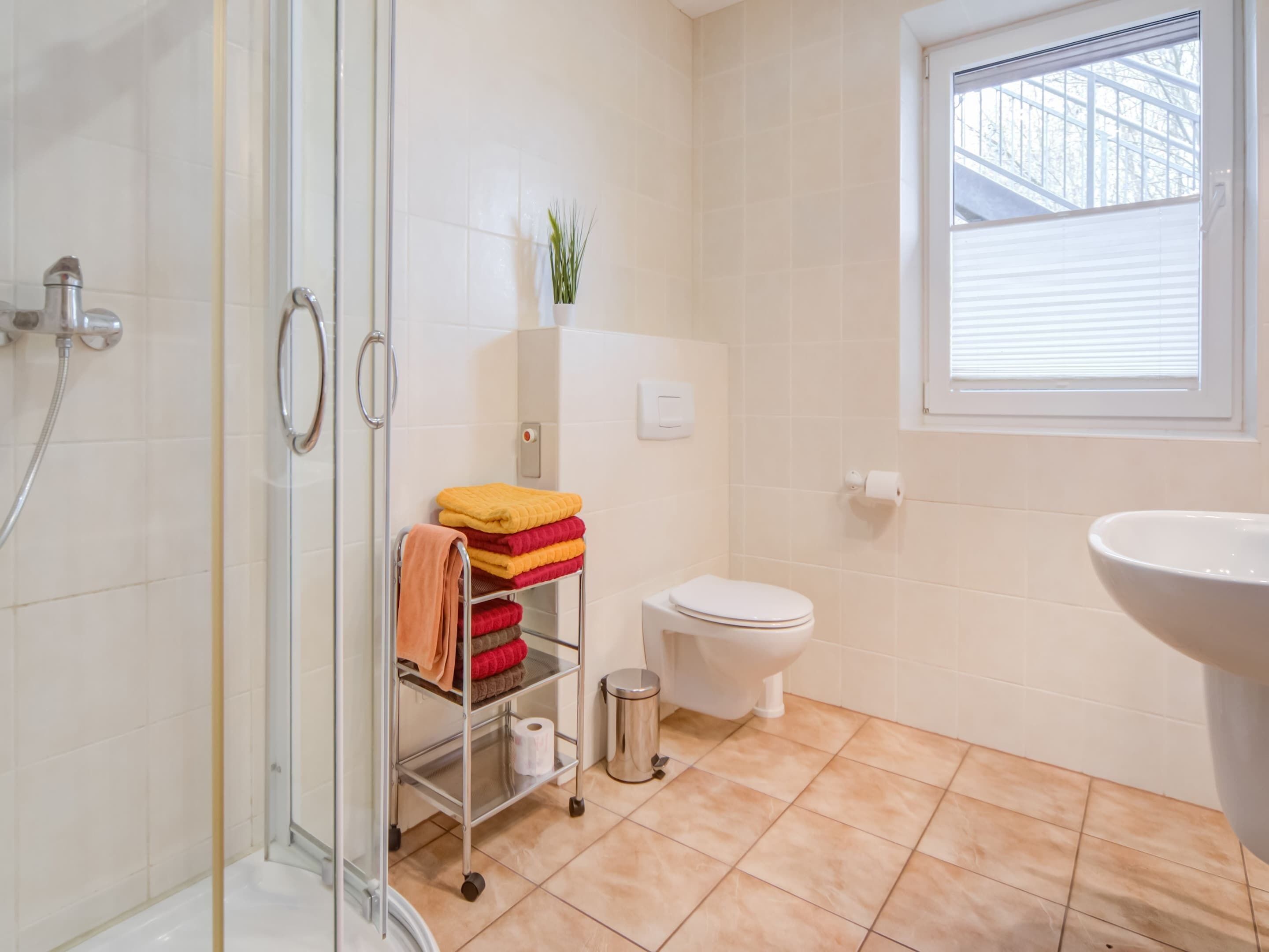A bathroom with a glass shower enclosure, a toilet, a sink, and a window with blinds.