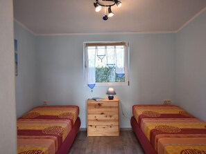 A room with two beds, a wooden dresser, a window with curtains, and a ceiling light fixture.