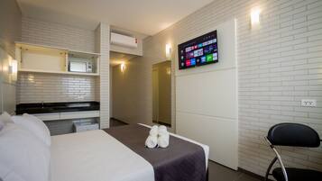 A hotel room with a bed, a TV mounted on the wall, a small kitchenette, and a chair.