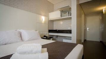 A modern hotel room with a large bed, a bedside table, a built-in TV, and a small kitchenette.