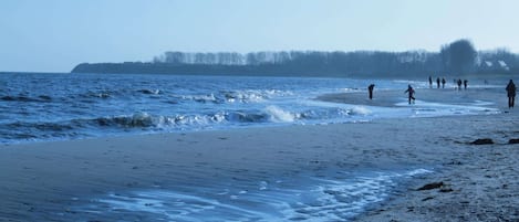 En strand med folk som promenerar längs strandkanten, vågor som slår in mot sanden och en avlägsen trädrad i horisonten.