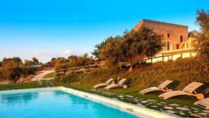 A swimming pool with lounge chairs, a building with a brick facade, and a hilly landscape. - Belvilla by OYO Corte Dorata (Scicli)