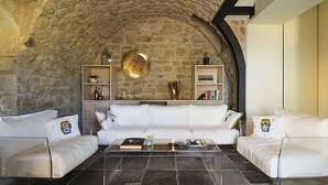 A modern living room with a stone wall, white leather sofas, a glass coffee table, and built-in shelves with decorative items. - Belvilla by OYO Corte Dorata (Scicli)