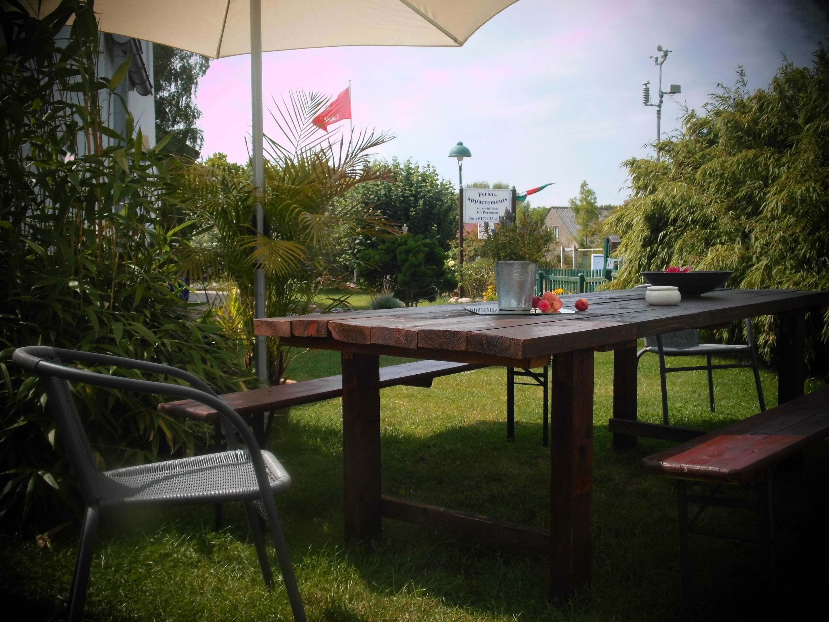 An outdoor dining area with a wooden table and benches, surrounded by lush greenery and a flag.