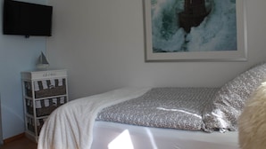 Un dormitorio con cama, televisor montado en la pared, estante con cestas y un cuadro con una ola.