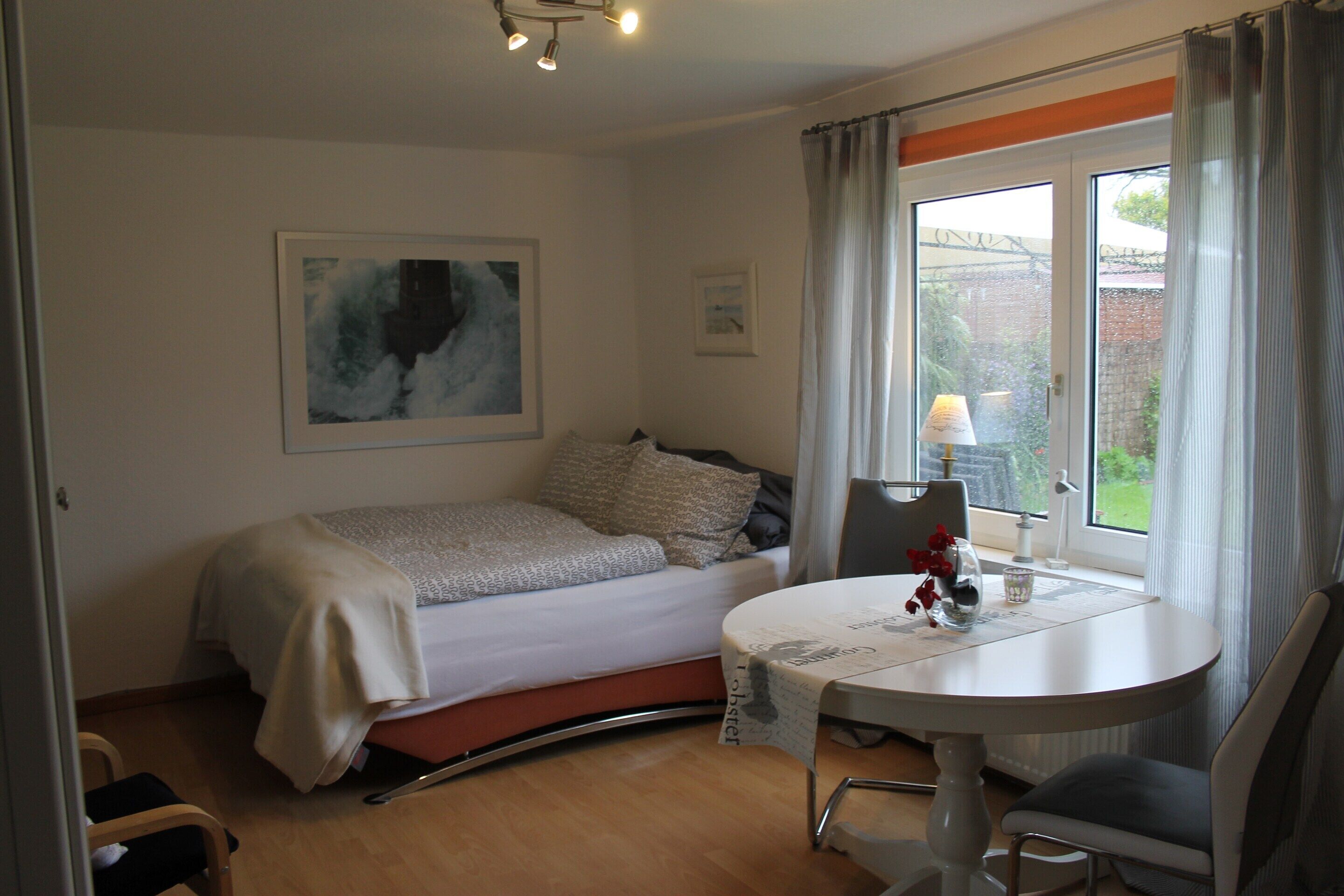 Quarto com cama, mesa de cabeceira com luminária, cadeira e janela com cortinas.