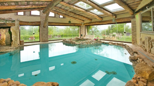 An indoor swimming pool with a rustic wooden ceiling, large windows, and a stone border. - Villa Solace - Comfort Charm (Guardea)