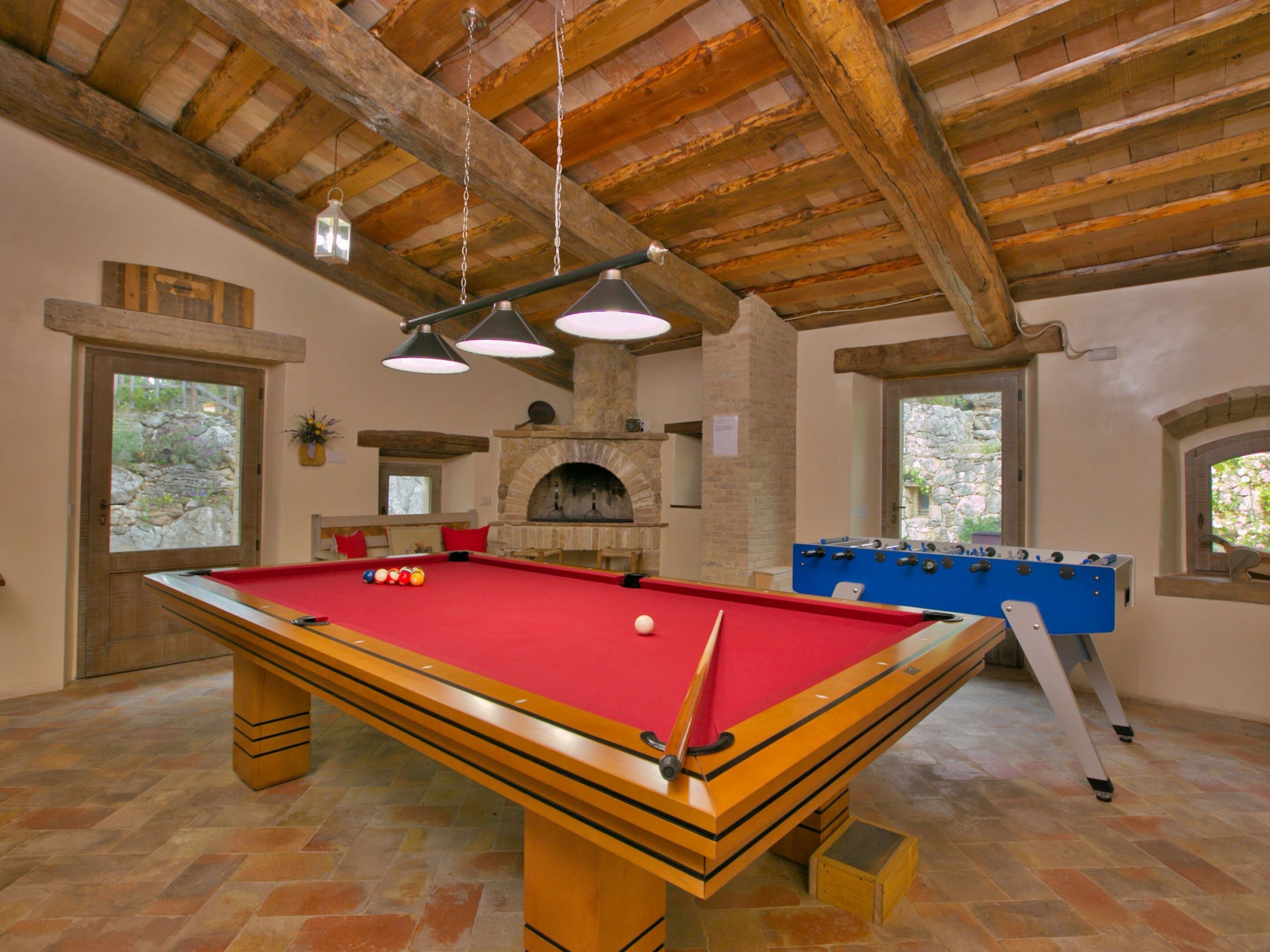 A pool table with a red felt surface, a foosball table, and a stone fireplace.