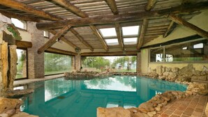 An indoor swimming pool with a stone border, wooden beams, and large windows. - Villa Solace - Comfort Charm (Guardea)