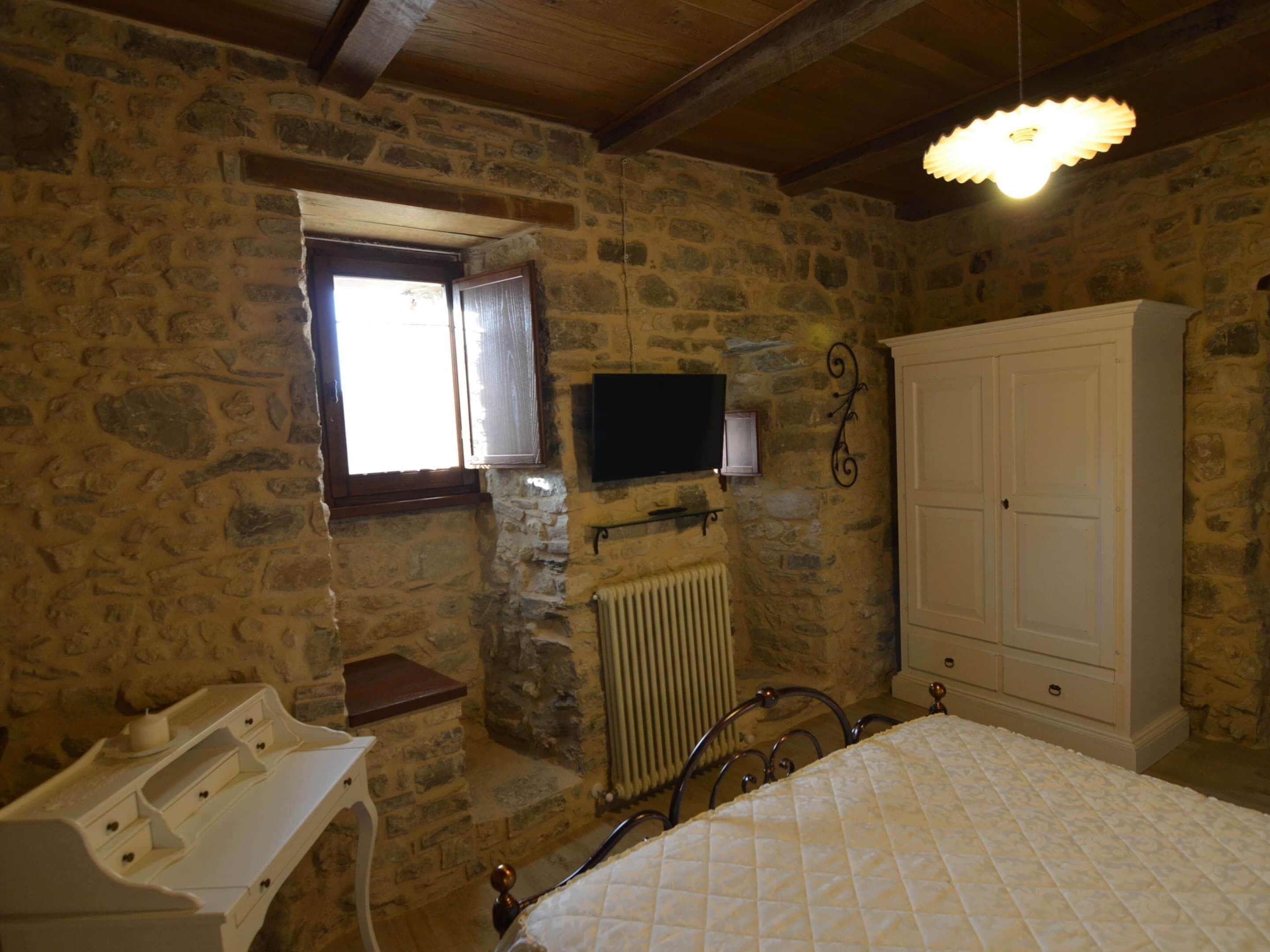 Ein Schlafzimmer mit Steinwänden, ausgestattet mit einem Bett, einem Schminktisch, einem Heizkörper, einem an der Wand montierten Fernseher und einem Kleiderschrank.