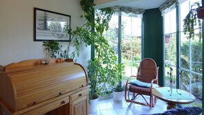 Una habitación con un armario de madera, una silla de mimbre, una mesa y un ventanal con cortinas.