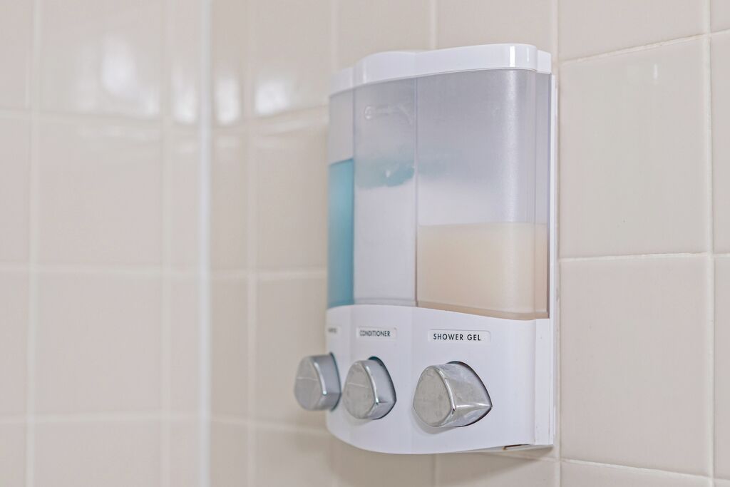 A shower gel dispenser with a transparent bottle and a pump, mounted on a tiled wall.