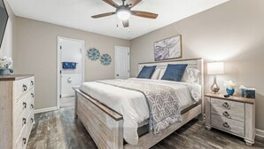 Une chambre à coucher comprenant un lit, une table de chevet, une commode, un ventilateur de plafond et une porte.