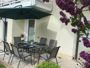An outdoor patio with a table and chairs, a green umbrella, and purple flowers.
