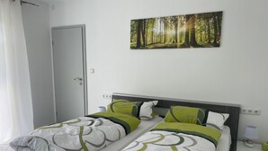 Una cama doble con ropa de cama verde y blanca, una mesita de noche con lámpara y un cuadro con motivos forestales en la pared.