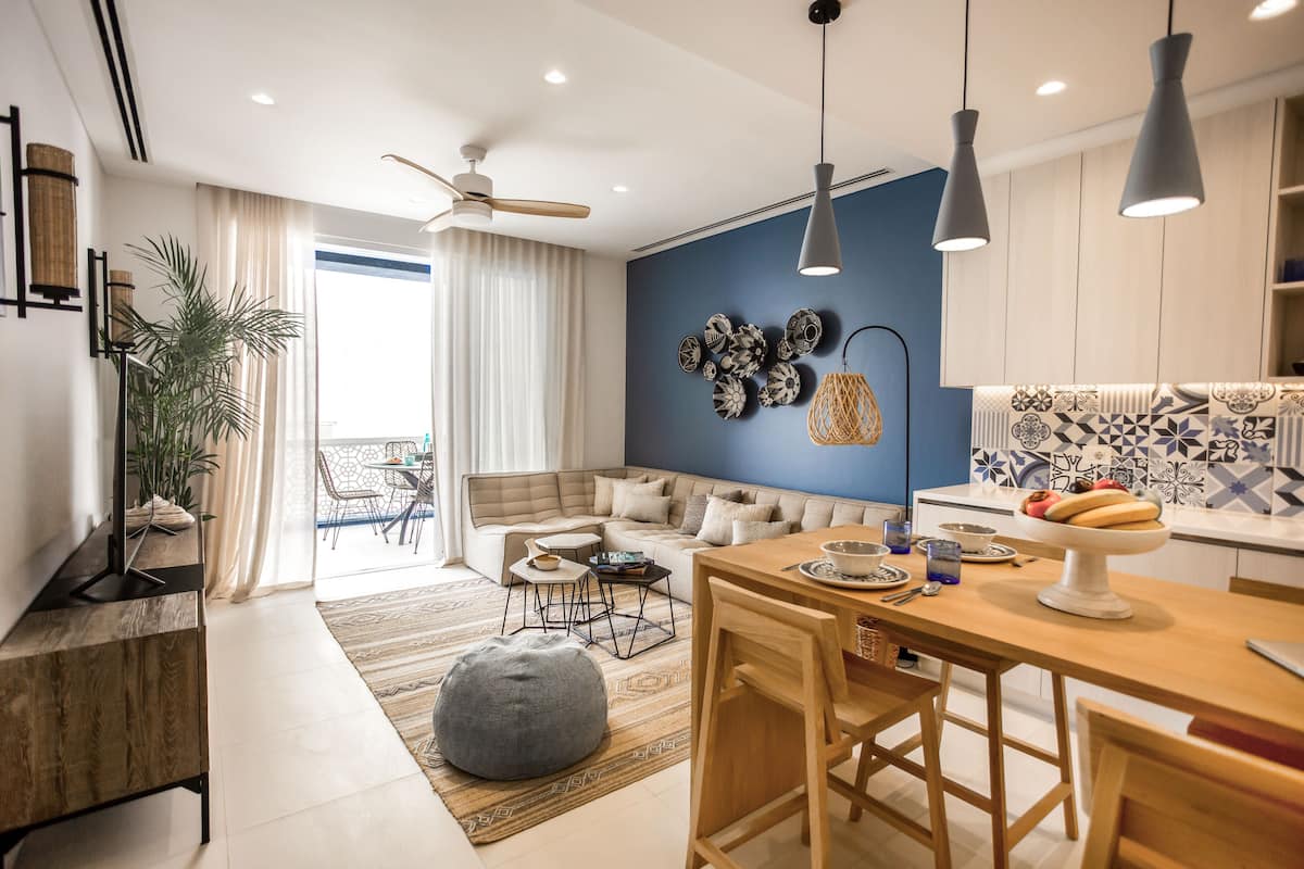 A modern living room with a sofa, coffee table, and dining area. There is a ceiling fan, pendant lights, and a large window with curtains.