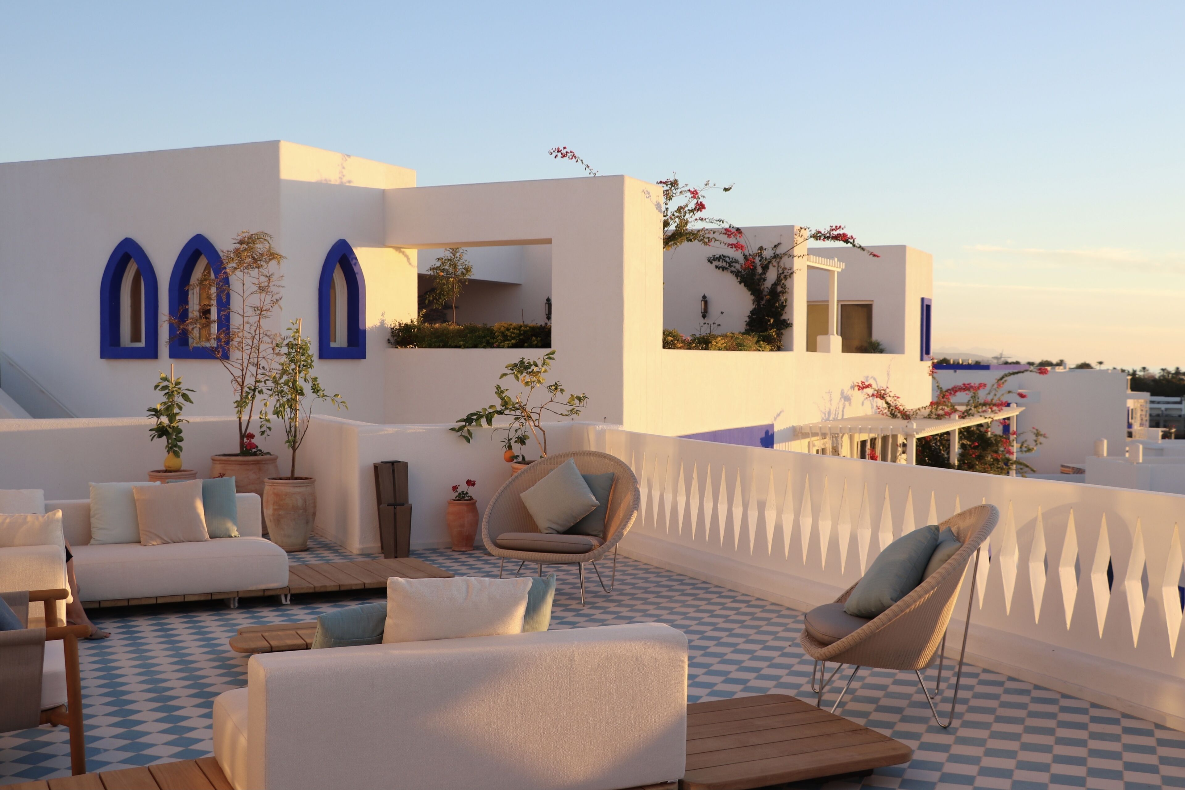 Una terraza en la azotea con suelo a cuadros, paredes blancas, ventanas con marcos azules y mobiliario exterior.