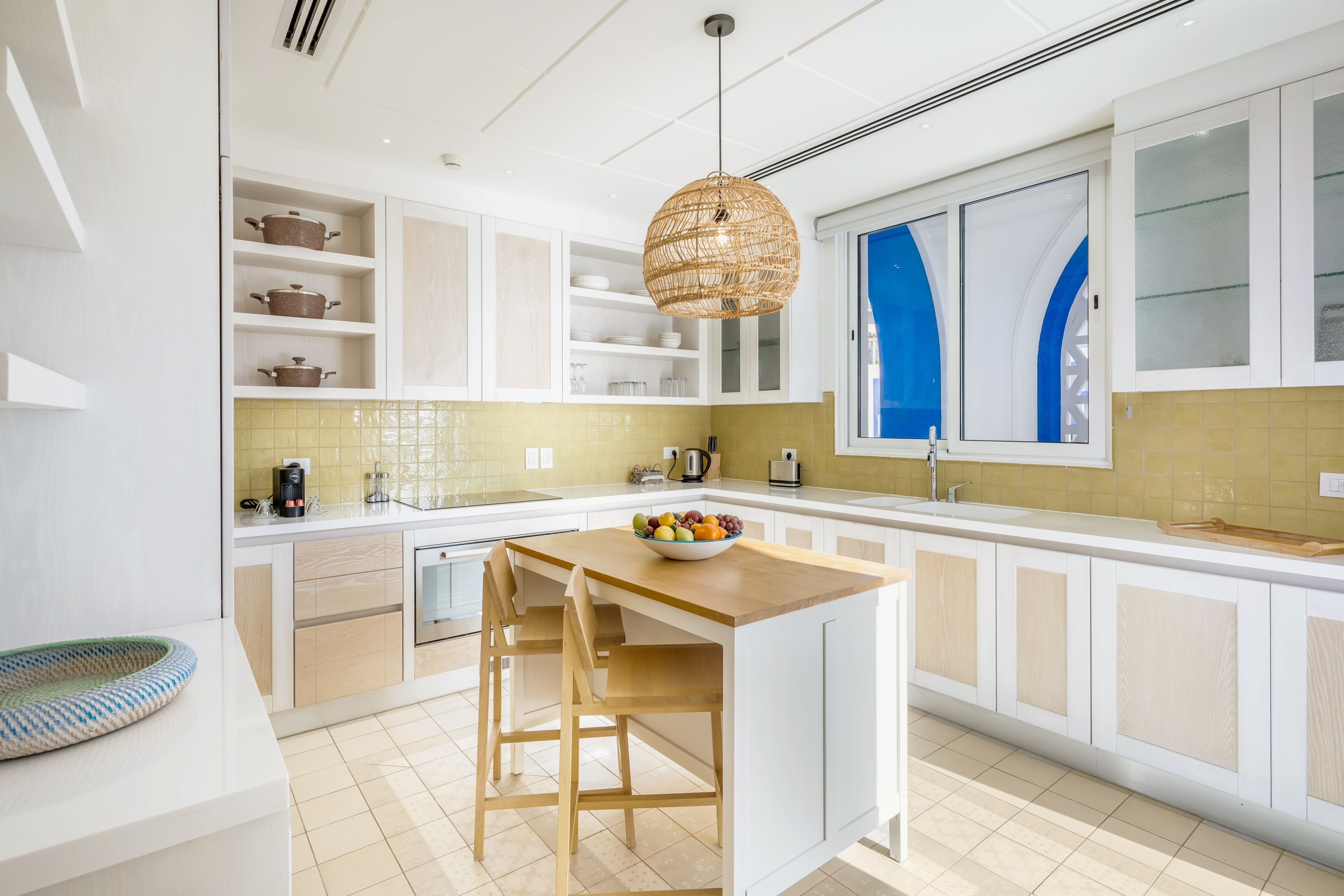 Una cocina moderna con armarios blancos, una isla central con taburetes, una lámpara colgante y un ventanal con cortinas azules.