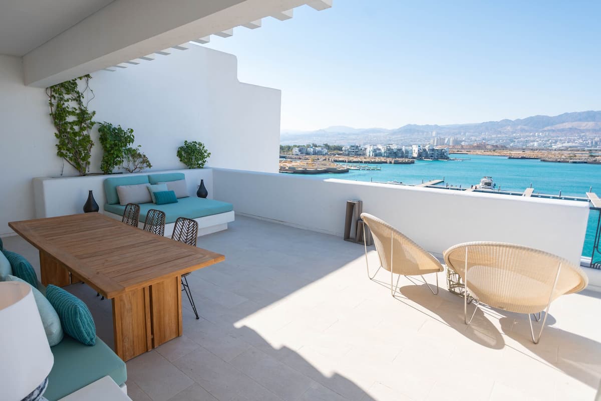 A balcony with a wooden table and chairs, a sofa with cushions, and a view of the sea and cityscape.