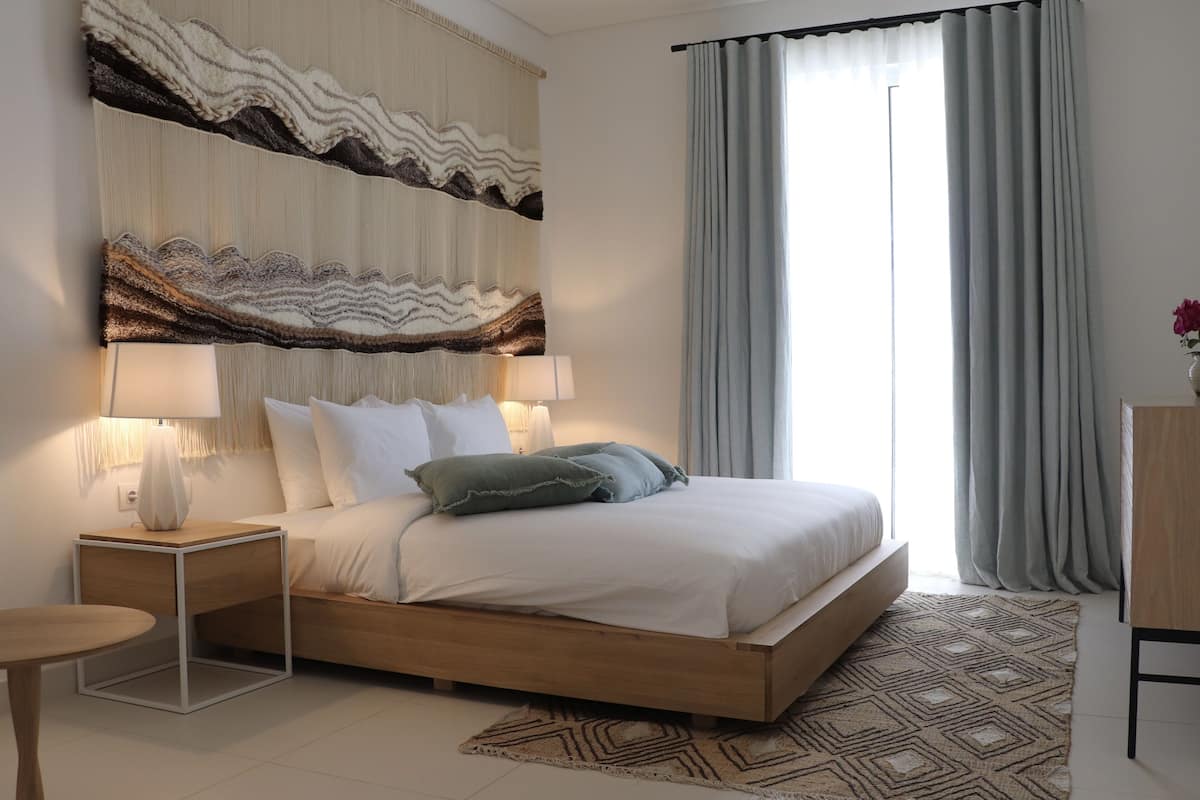 A modern bedroom with a wooden bed, two bedside tables, a large abstract wall hanging, and a view of the outdoors through sheer curtains.
