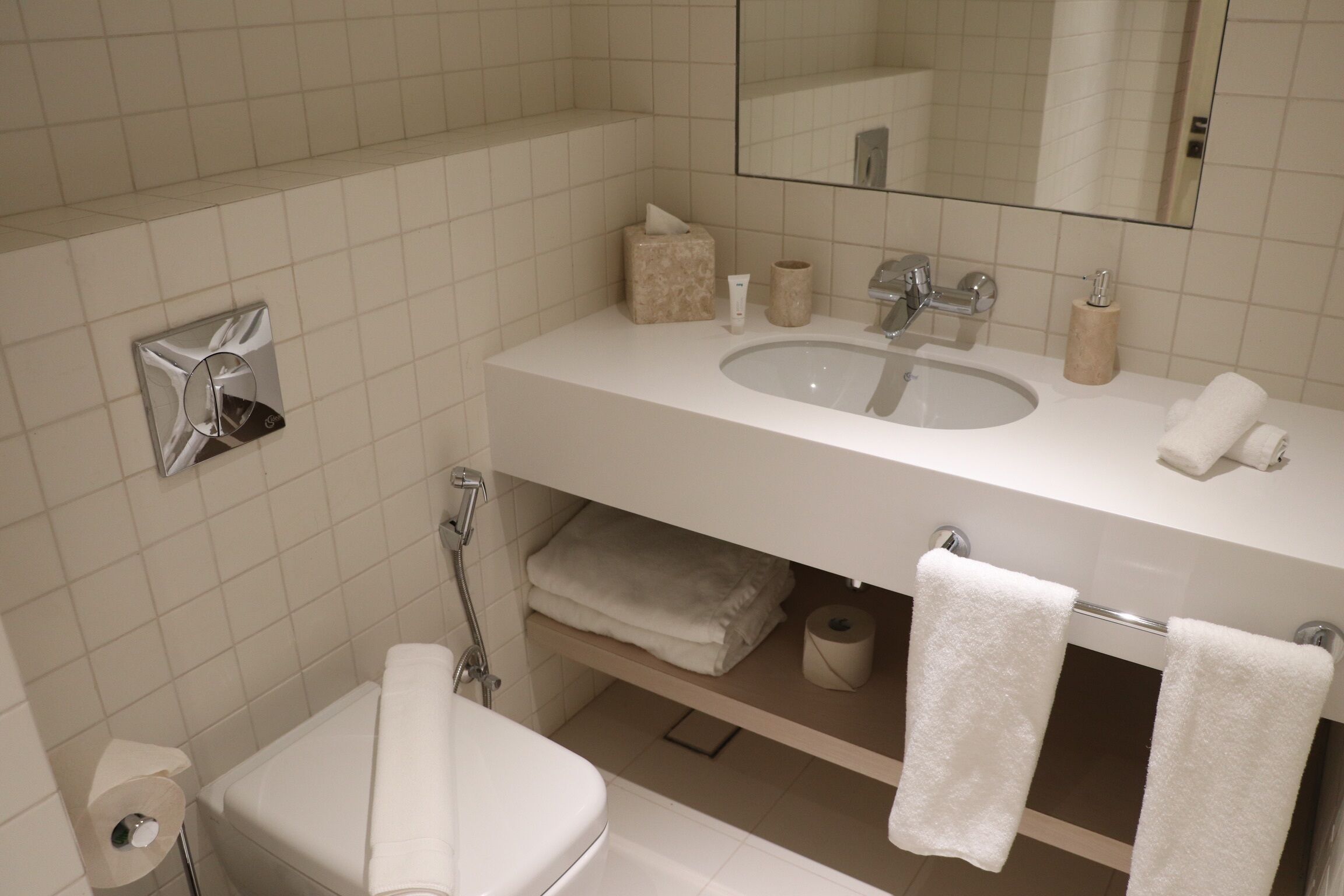 Une salle de bain avec un lavabo blanc, un miroir et un porte-serviettes. On y trouve également des toilettes avec un distributeur d’essuie-tout et un rouleau de papier toilette.