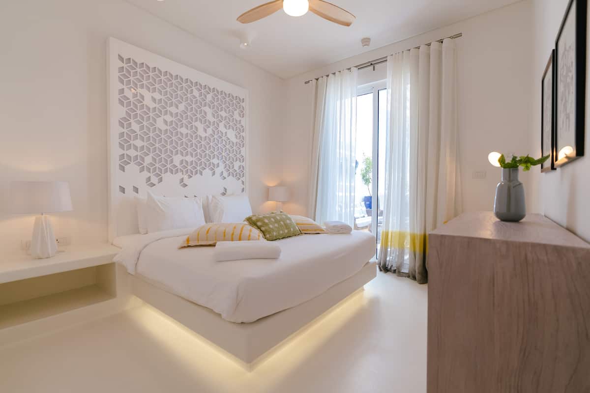 A modern bedroom with a large bed, a bedside table with a lamp, a decorative wall piece, and a view of the outdoors through a window with curtains.