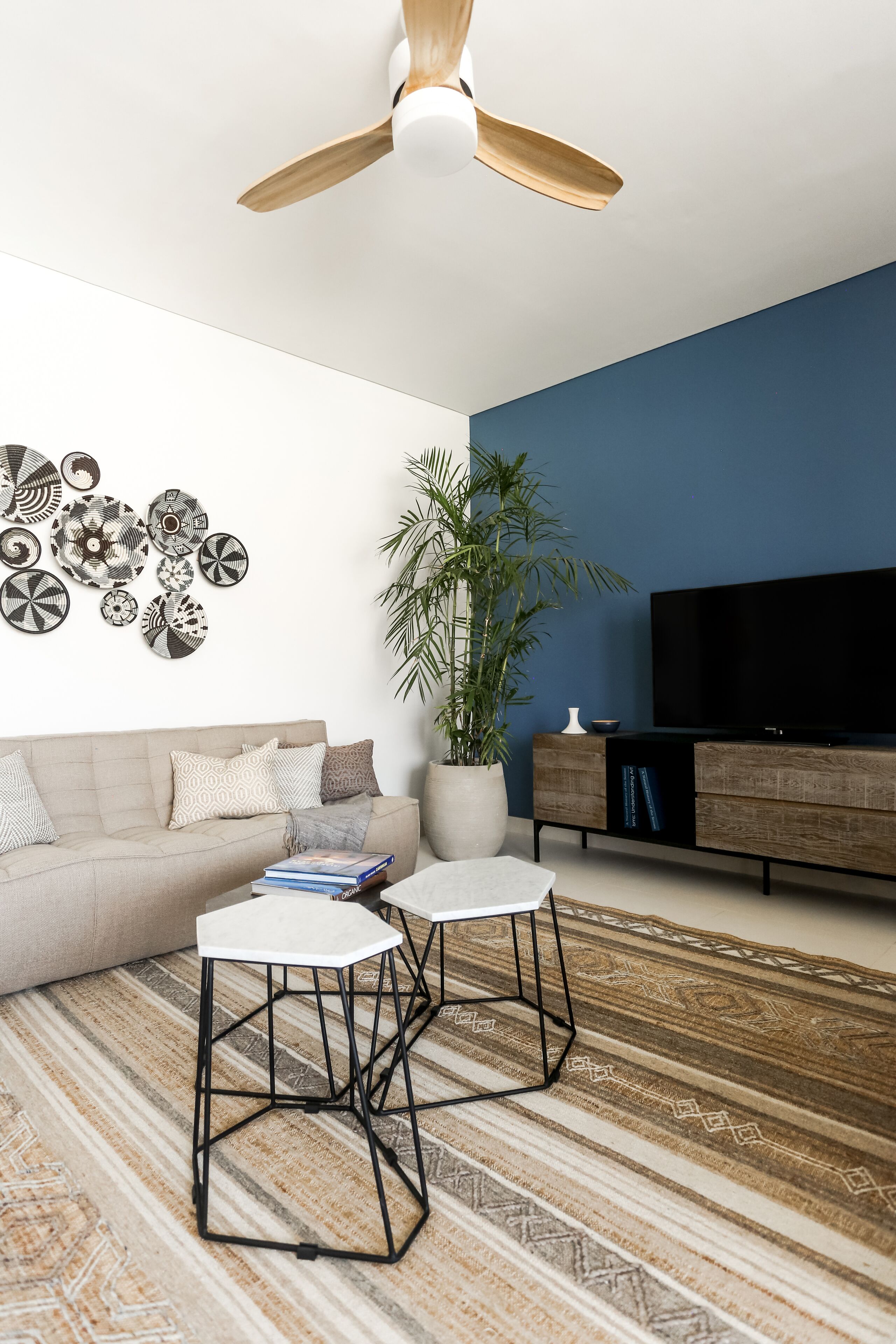 Ein modernes Wohnzimmer mit einer Couch, zwei weißen Tischen, einer großen Topfpflanze, einem Flachbildfernseher und einem Deckenventilator.